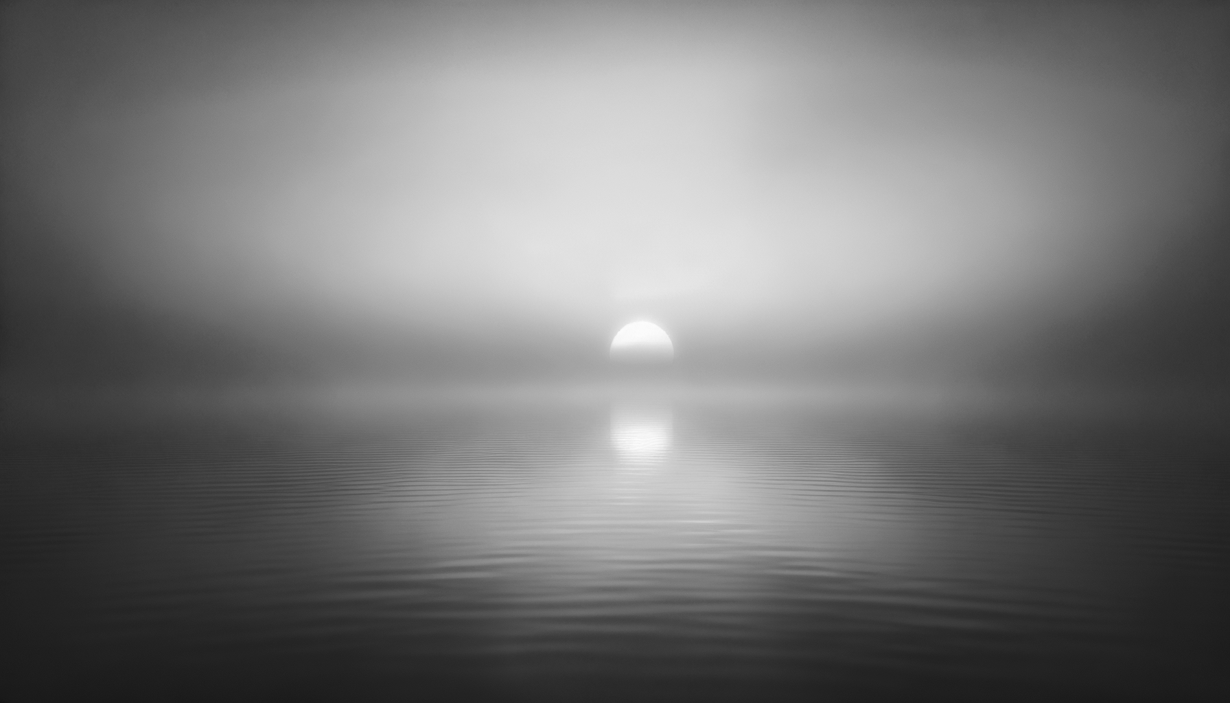 Black and white photograph of the sun setting over calm water, with gentle ripples and a reflective surface.
