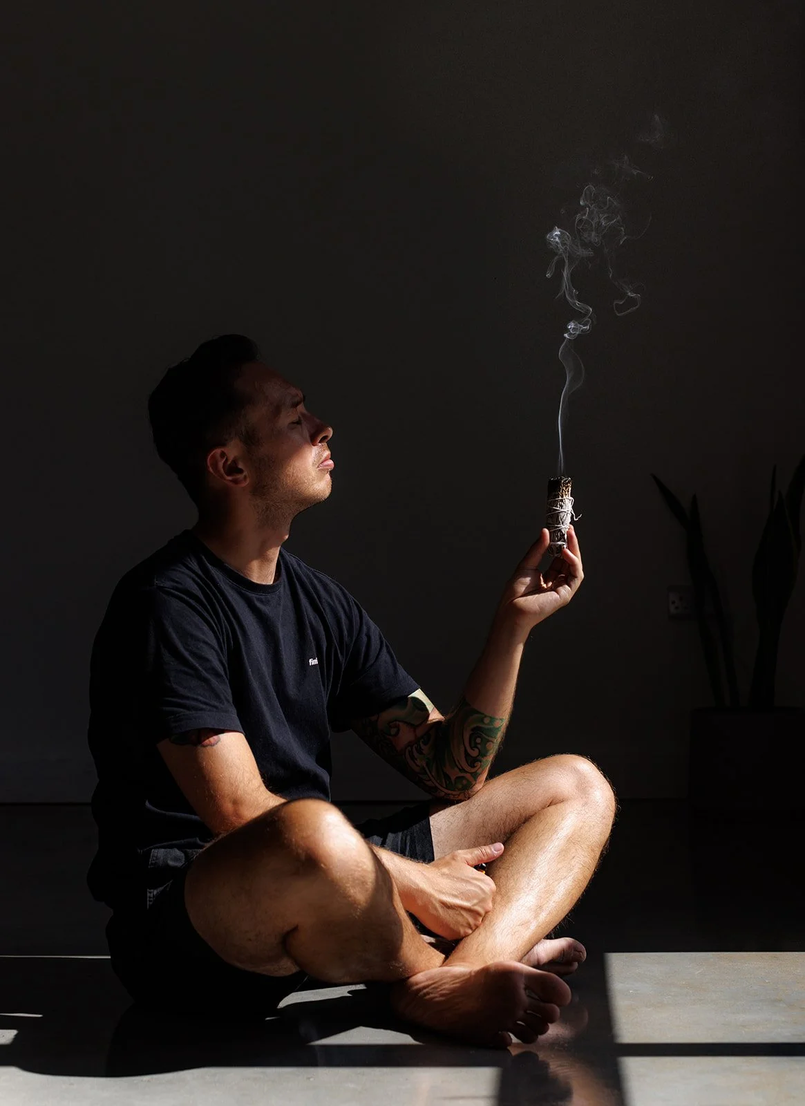 A man sitting cross-legged on the floor holding a lit sage, with smoke rising from it, in a dark room with minimal lighting.
