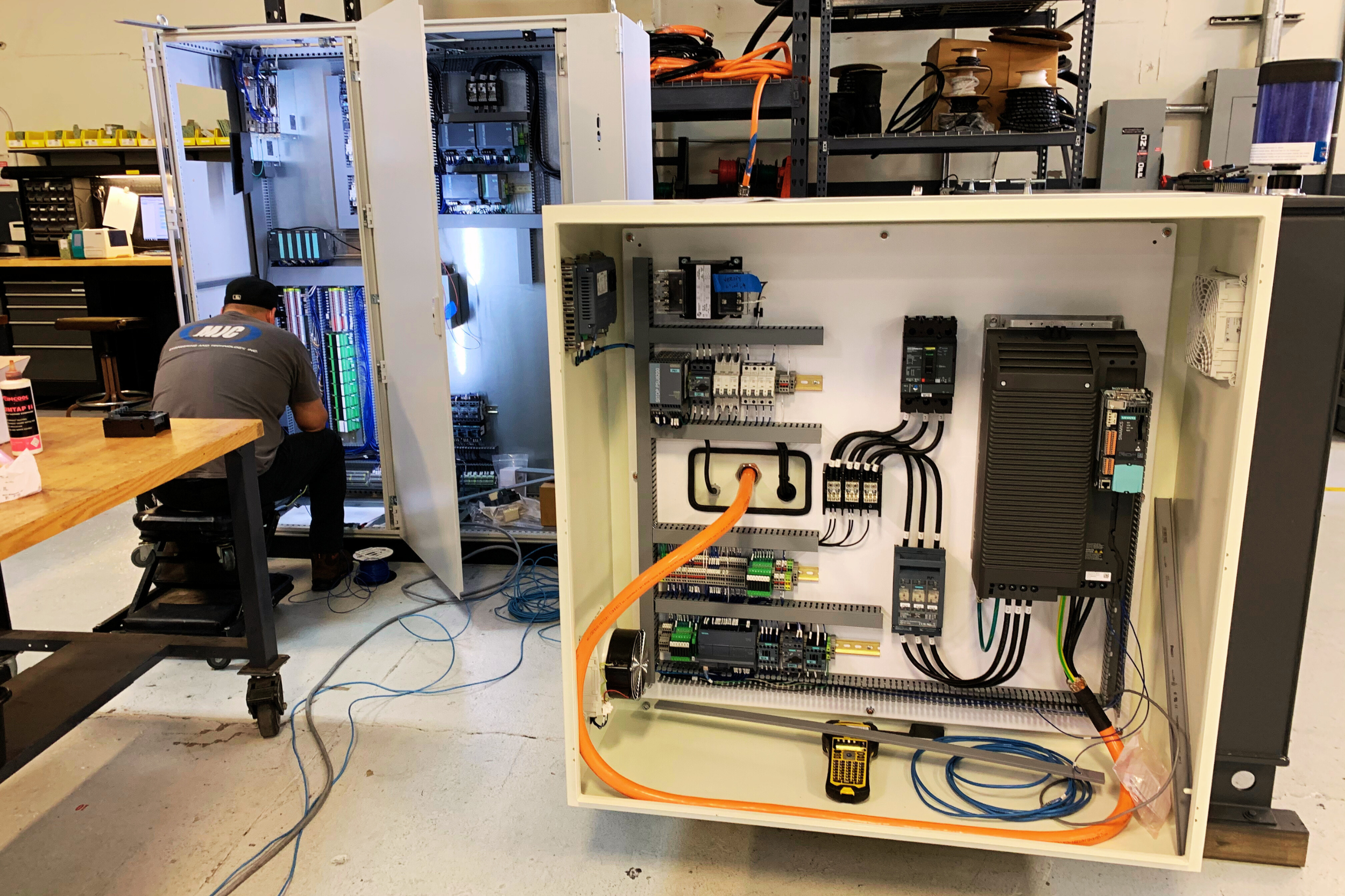 A technician repairing or assembling HVAC or industrial control systems inside a large white electrical control panel with various components and wiring. Another open electrical cabinet with more components is visible behind, with tools and equipment on shelves.