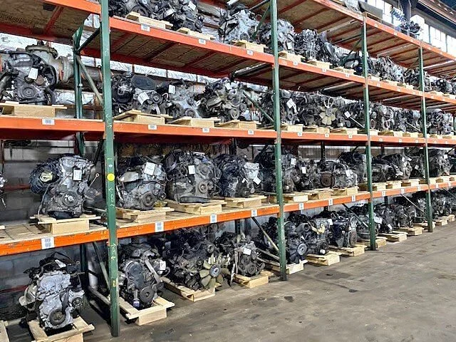 Multiple car engines stacked on industrial metal and wood shelving units in a warehouse.