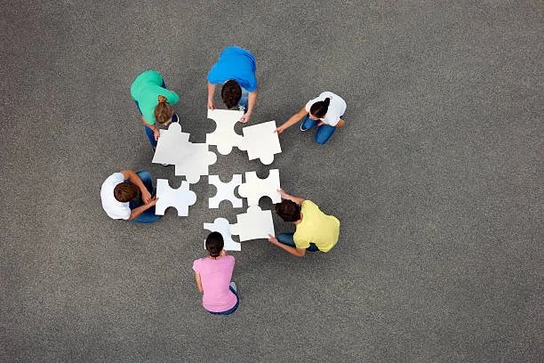 Six people assembling large white puzzle pieces on a gray surface, viewed from above.