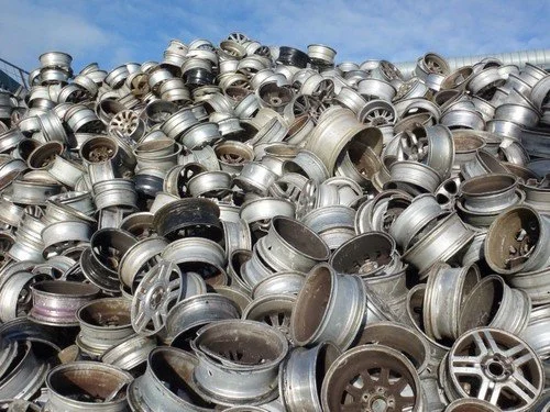 A large pile of aluminum car rims stacked outdoors against a blue sky.