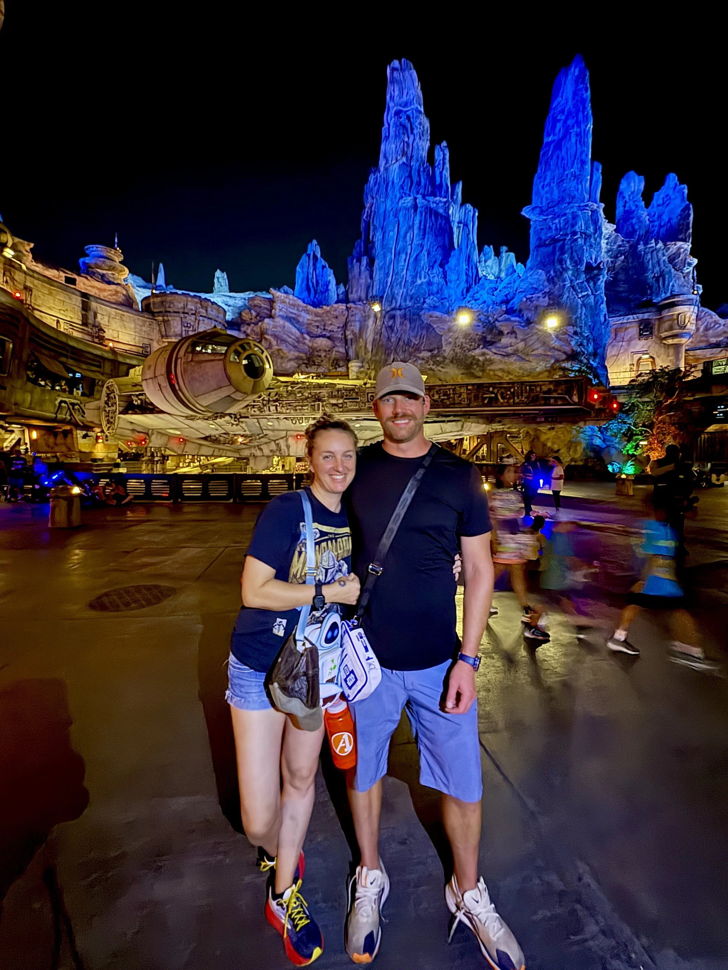 A couple standing together at night in front of a themed area with tall blue rock formations, likely at a theme park like Disneyland or Disney California Adventure, with glowing lights and other visitors walking around.