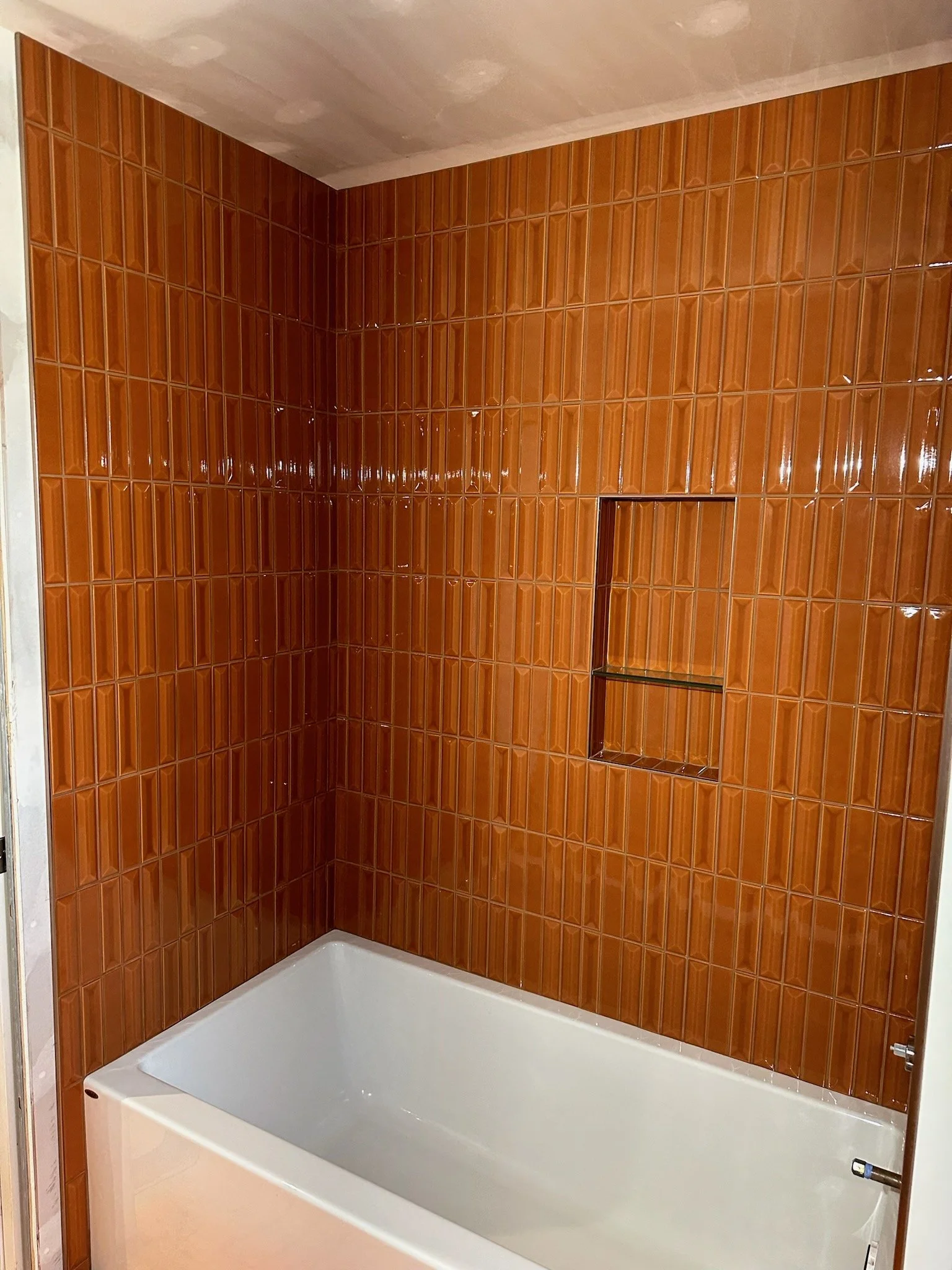 A shower area with orange-brown ceramic tile walls, a built-in shelf, and a white bathtub.