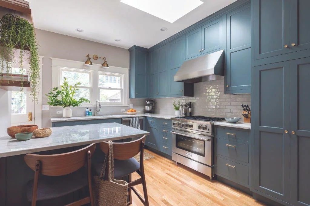 Modern kitchen with blue cabinets, stainless steel stove and range hood, white subway tile backsplash, skylight, window with plants, and wooden flooring.