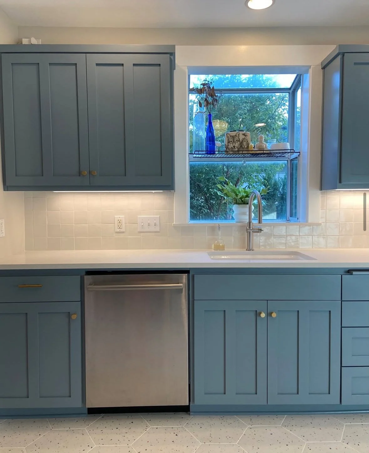 Kitchen with blue cabinets, white countertop, white tile backsplash, stainless steel dishwasher, and a window with decorative items and plants.