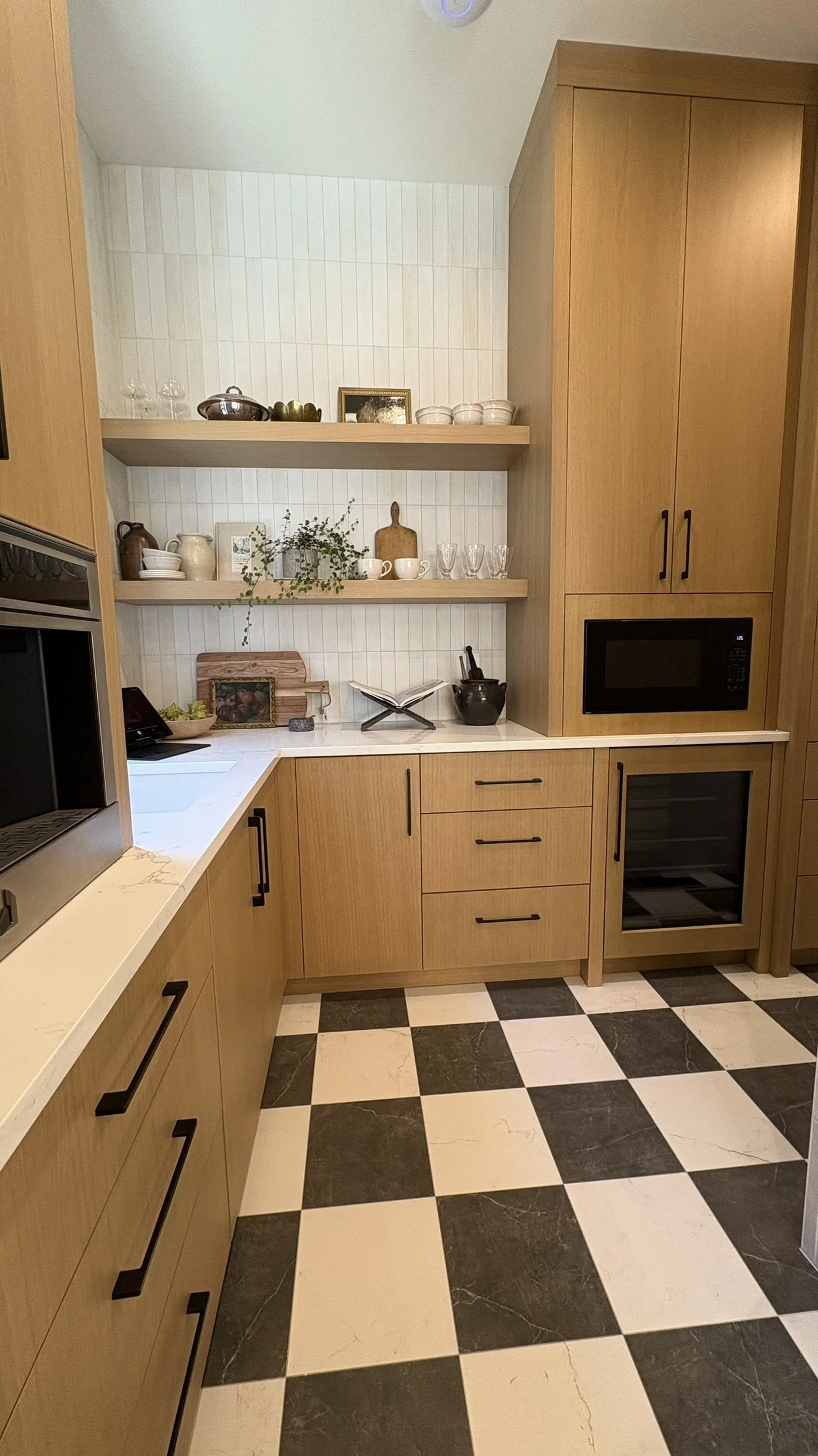 Kitchen with wooden cabinets, black handles, black and white checkered floor, built-in microwave, wine fridge, open shelves with decorative items, and white countertop.