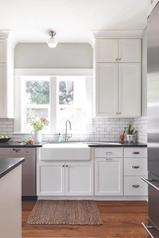 Bright kitchen with white cabinets, white subway tile backsplash, and a large window above the sink, with a brown rug on wooden floor.