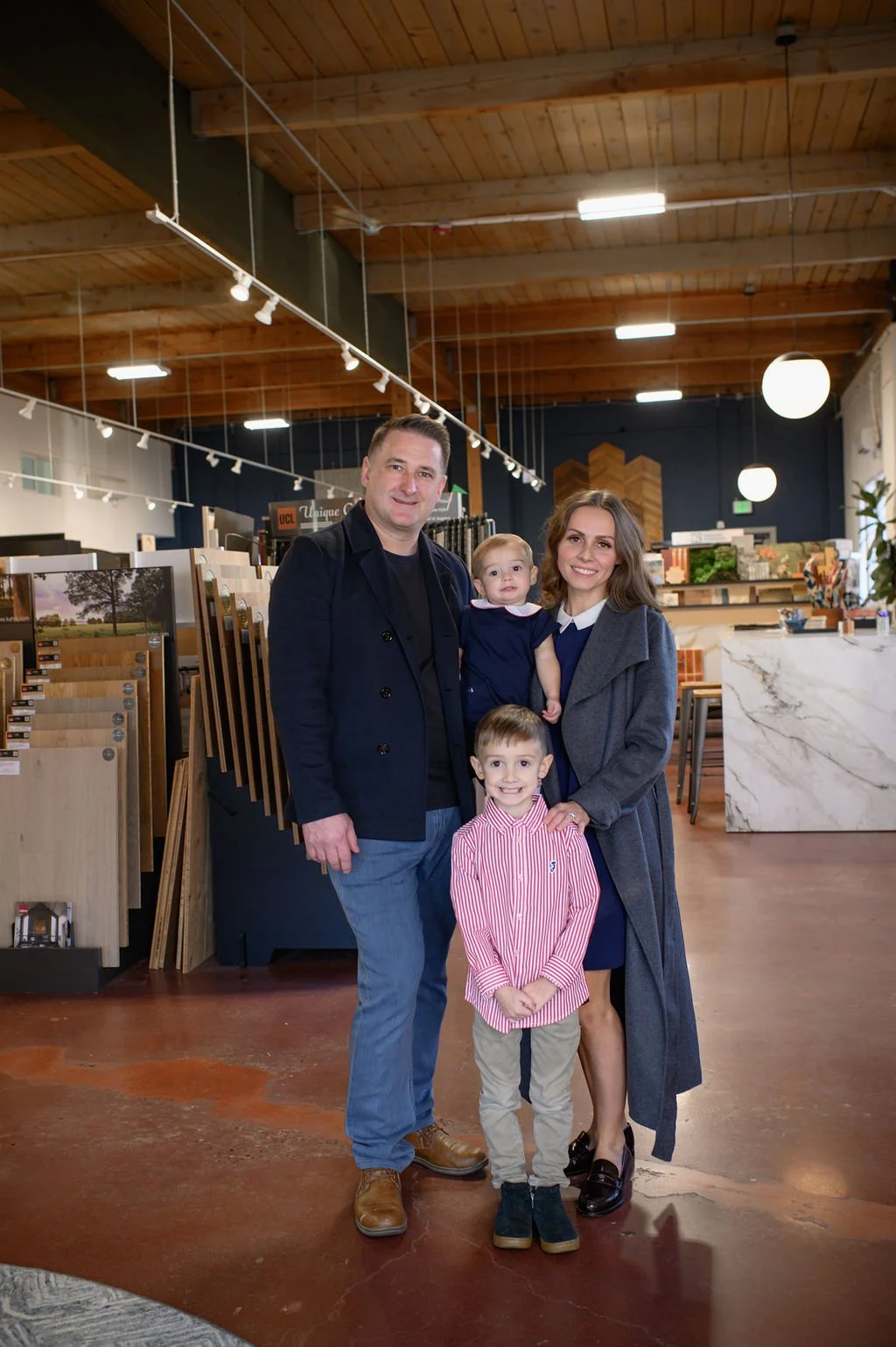 A family of four standing inside a modern store with wood and marble decor. The man and woman are smiling, with two children, a boy and a girl, standing in front and to the side of them.