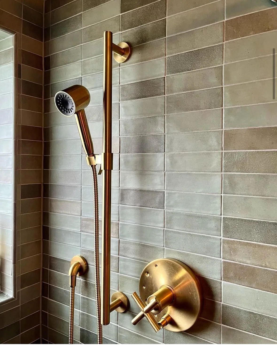 A modern shower with a yellow gold finish, including a handheld showerhead, an adjustable vertical shower bar, and a round control panel with a lever handle. The wall is tiled with rectangular, earthy-toned tiles.