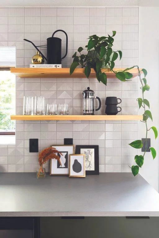 Kitchen shelf with a black kettle, a black coffee dripper, a French press, several glasses, potted plant, and framed art on a tiled wall.