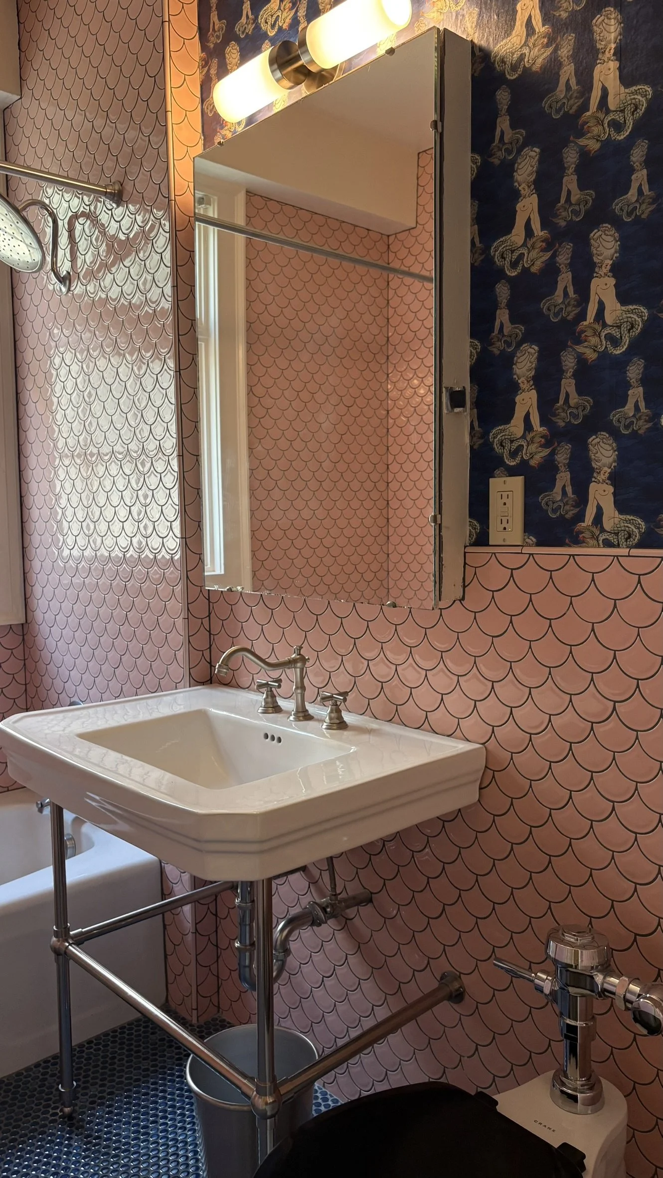 Bathroom with pink scalloped wallpaper, a white vintage sink with a metal frame, a mirror, a wall-mounted light fixture, and a partial view of a bathtub and plumbing fixtures.