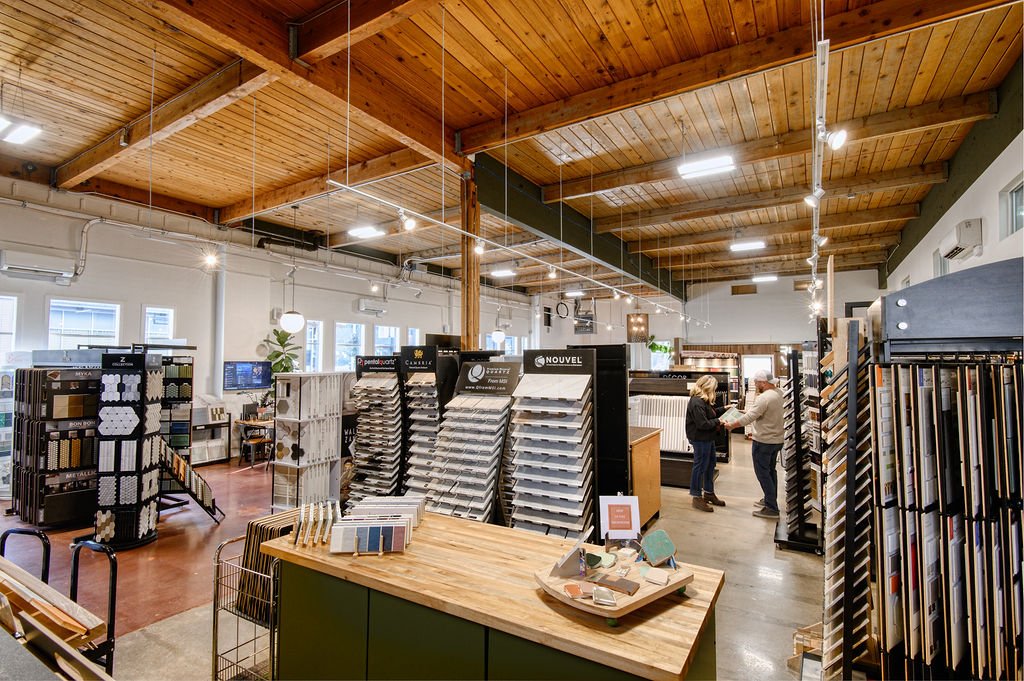 Interior of a flooring or home improvement store with displays of flooring samples, customers talking to staff, and wooden ceiling with industrial lighting.
