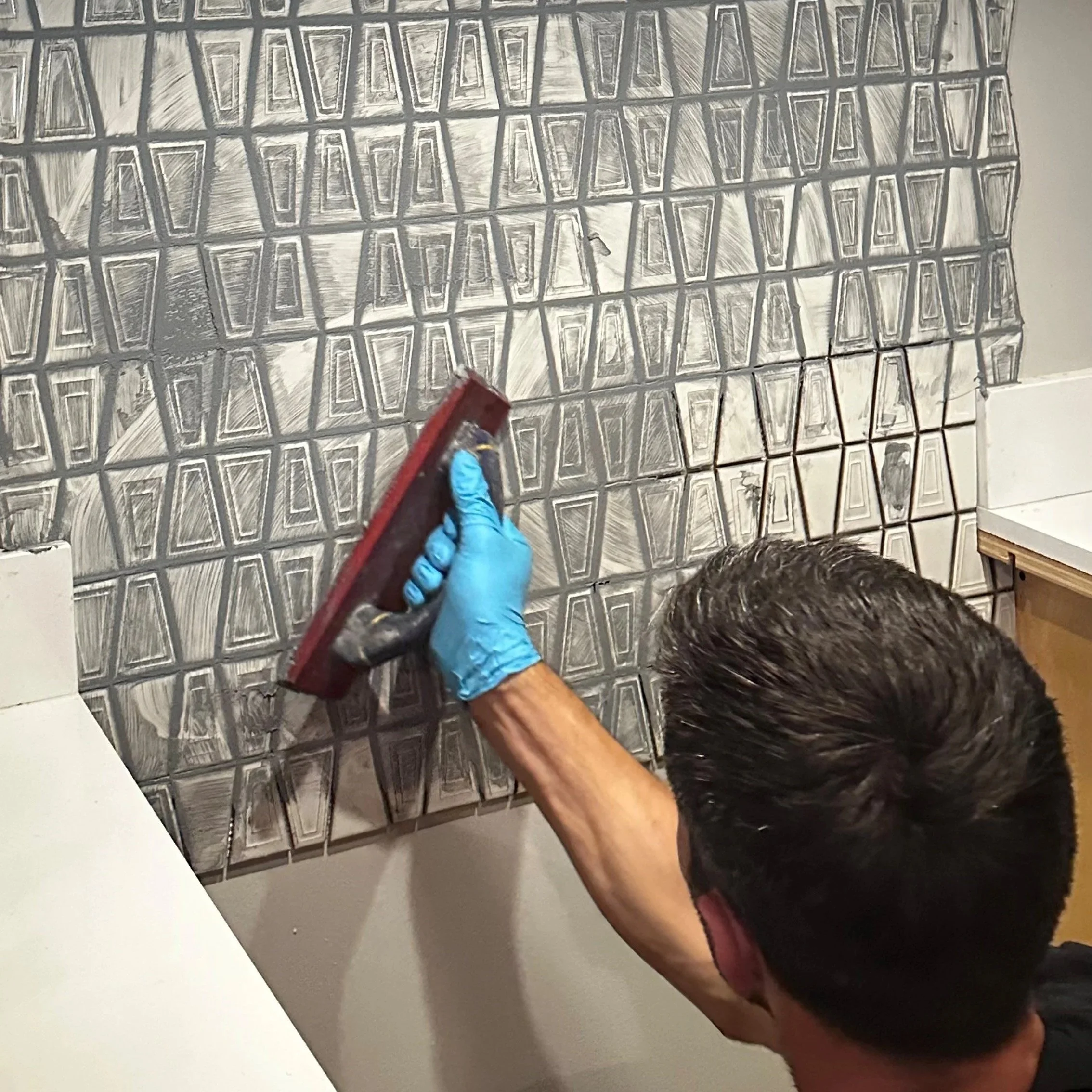 A person with short dark hair wearing a blue glove is using a scrubbing tool to clean or prepare a tiled wall with geometric pattern tiles.