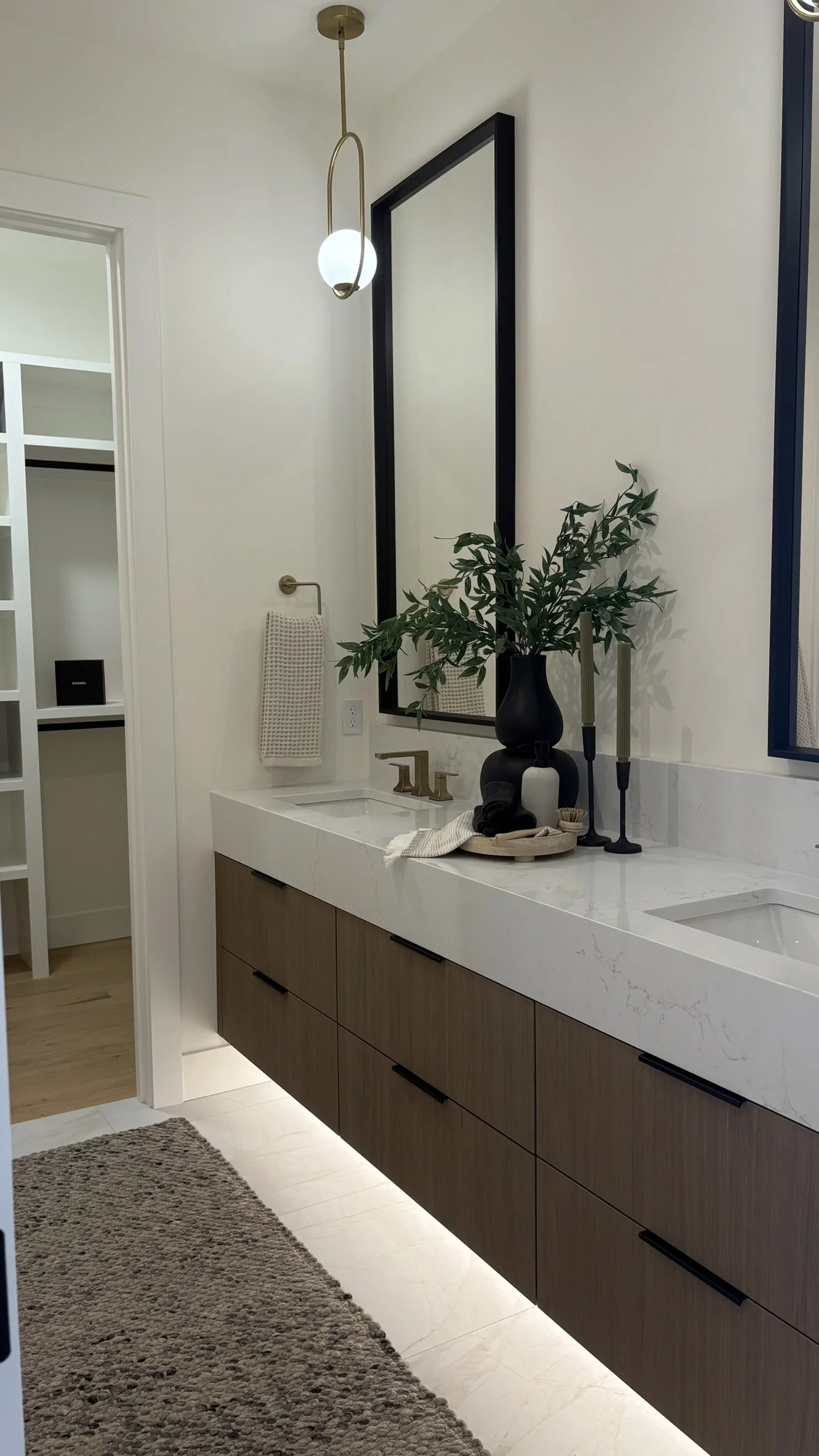 Modern bathroom with a white marble vanity, a large mirror, a black vase with greenery, brass fixtures, and black candlesticks, with a textured rug on the floor.