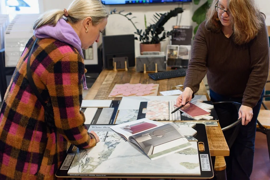 Two women are looking at design samples and color swatches on a table in a design studio or office.