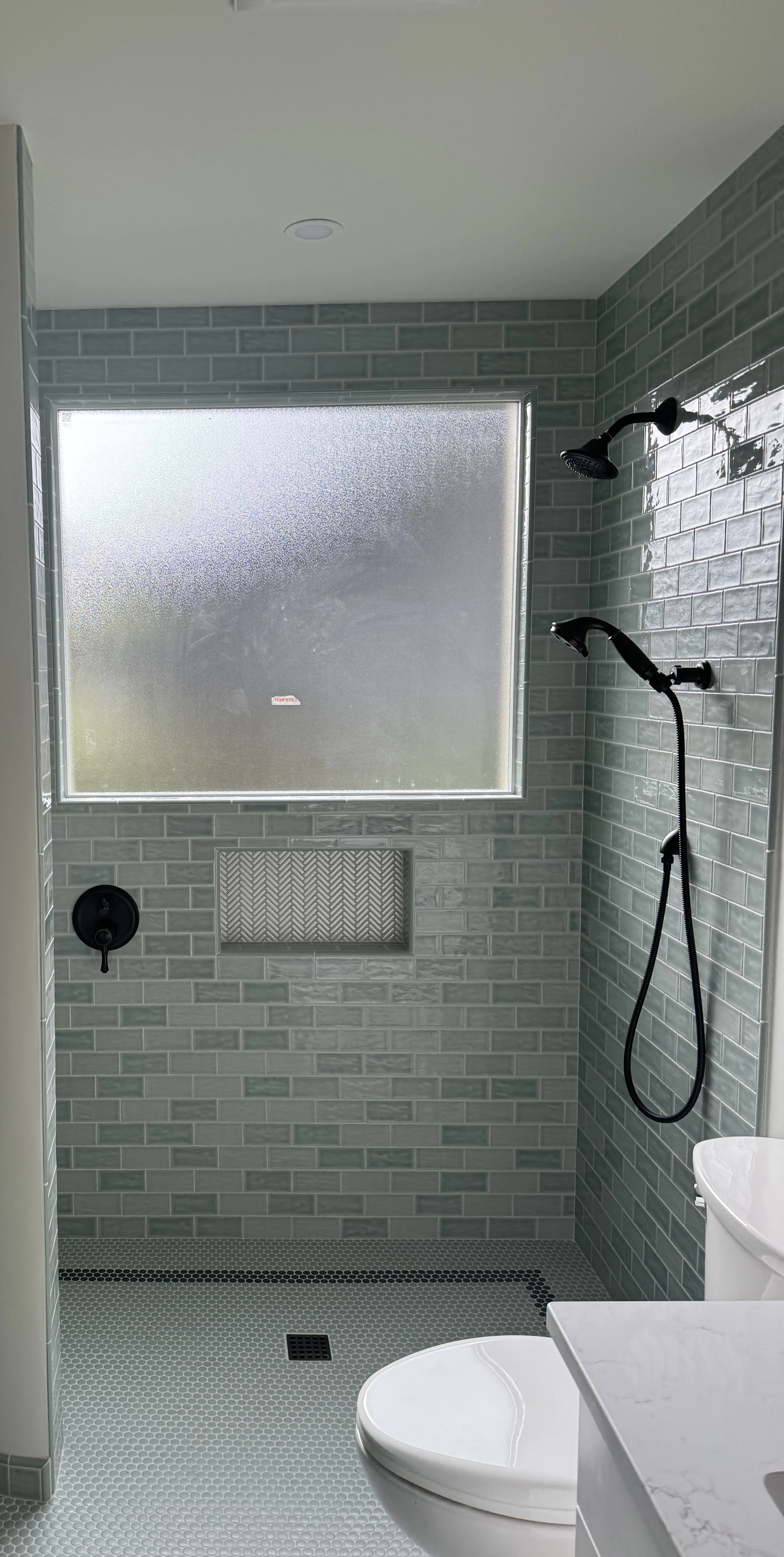 Modern bathroom shower with frosted window, gray subway tile walls, black shower fixtures, and a white toilet.