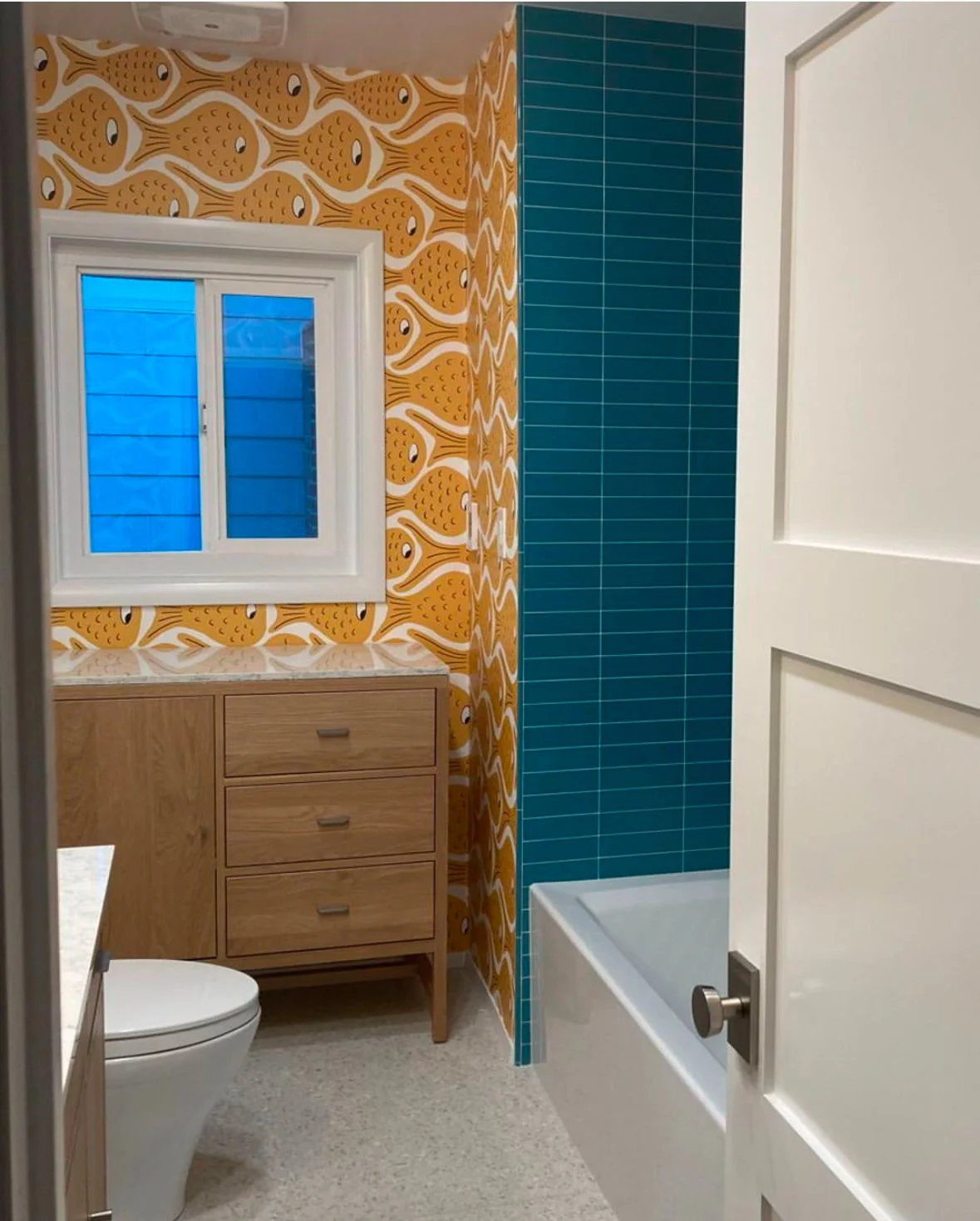 A bathroom with colorful wallpaper featuring yellow fish, a window with blue shutters, a wooden vanity, a toilet, and a bathtub with a blue tiled wall.