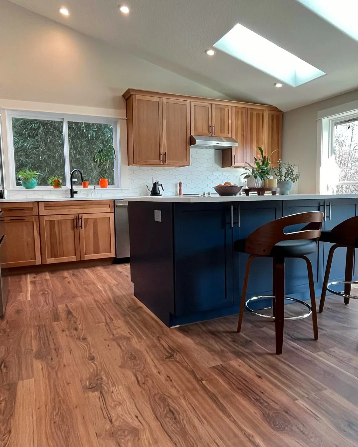 A modern kitchen featuring wooden cabinetry, a white hexagon tile backsplash, a kitchen island painted in dark blue, a skylight, and wooden floor.