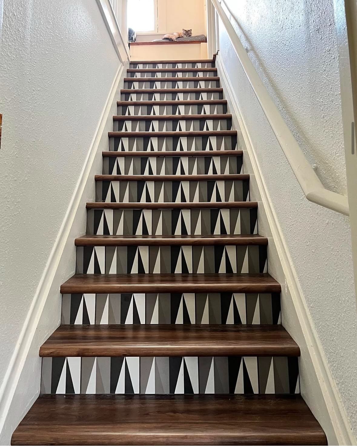 Indoor staircase with dark wood steps, white risers decorated with black and white geometric triangle pattern, white walls, and a kitten resting on a windowsill at the top of the stairs.
