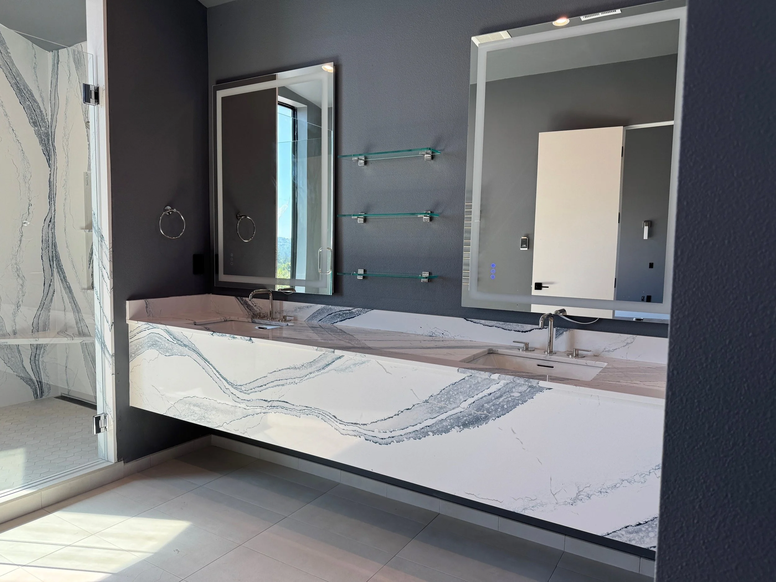 Modern bathroom vanity with two sinks, marble countertop, and two mirrors with built-in lighting. Grey walls, glass shelves, and a shower area with a glass door are visible.