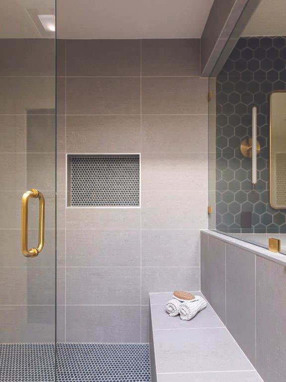 Modern bathroom shower area with glass door and gold handle, gray tiles, and a small bench with towels and a soap.