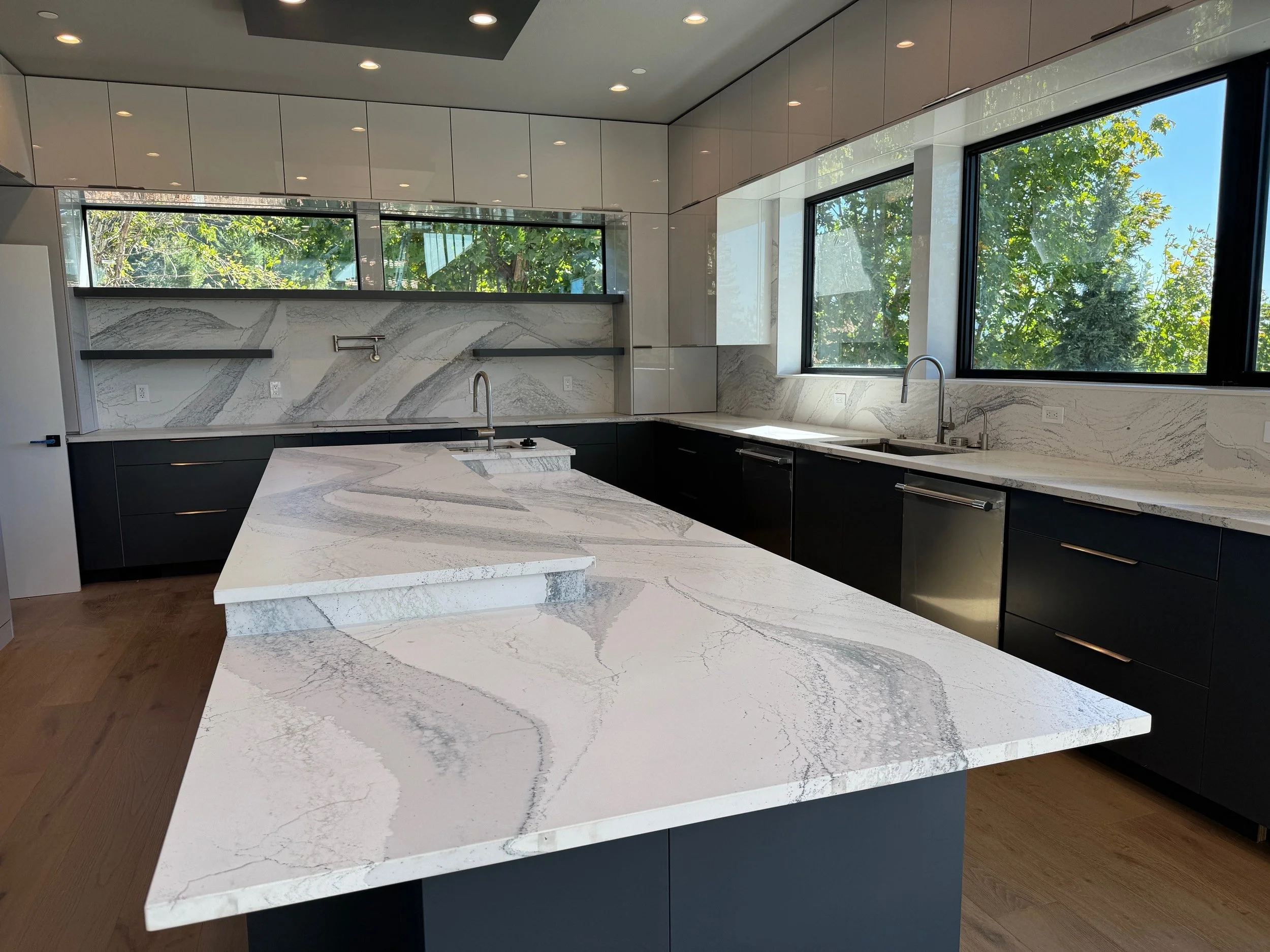 Modern kitchen with white marble countertops and backsplash, black lower cabinets, and large windows showing green trees outside.