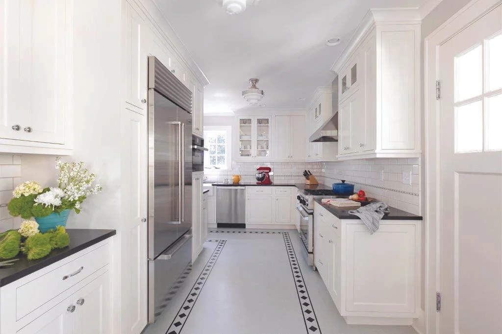 Bright white kitchen with black countertops, stainless steel appliances, and a decorative black and white tile floor border. There is a flower arrangement on the left counter, a red stand mixer, and a blue pot on the right stove. The room has natural