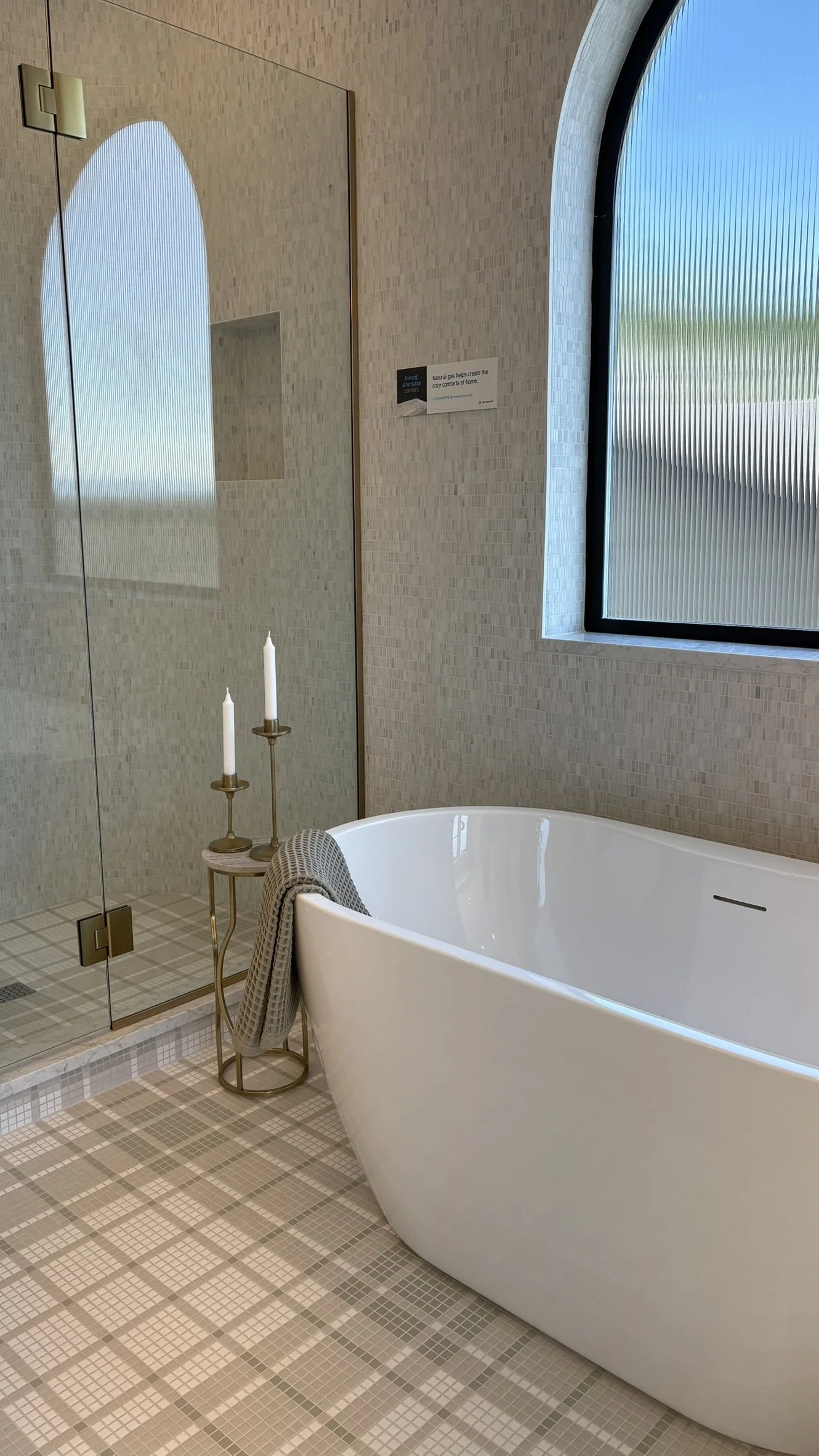 A modern bathroom featuring a freestanding bathtub, a glass shower enclosure, and a small side table with two white candles. There is a textured beige tiled wall and floor, a large arched window with frosted glass, and a mirror reflecting part of the
