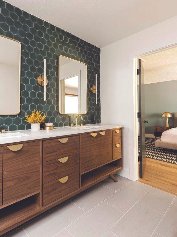 Modern bathroom vanity with a white countertop, two mirrors with gold frames, and black hexagonal tile wall behind it. There are gold and black wall-mounted light fixtures on either side of the mirrors, a potted plant on the vanity, and a doorway to a bedroom with a bed, nightstand, and lamp visible in the background.