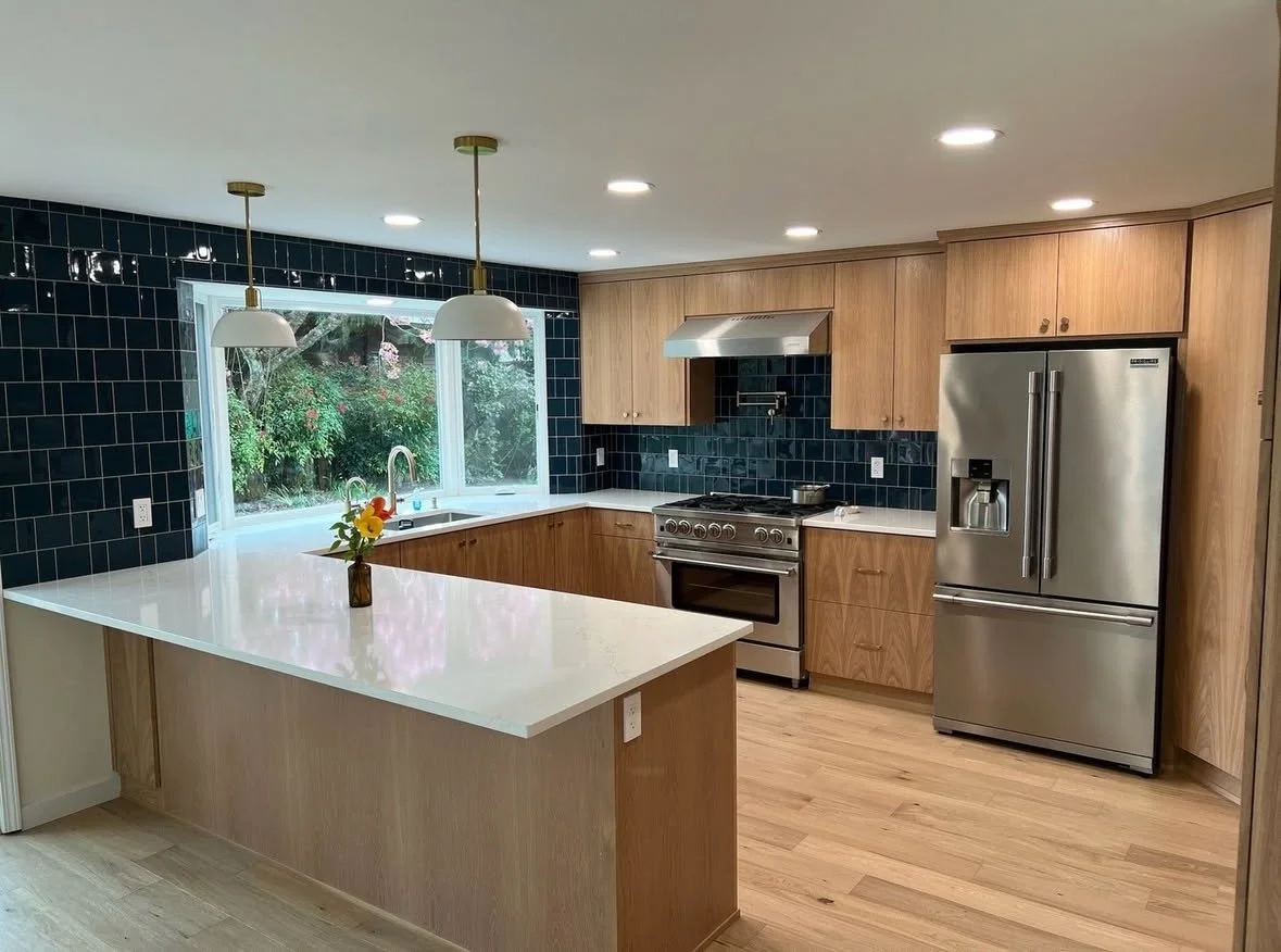 Modern kitchen with wooden cabinets, stainless steel refrigerator, stove, and dark blue tiled wall near a large window.