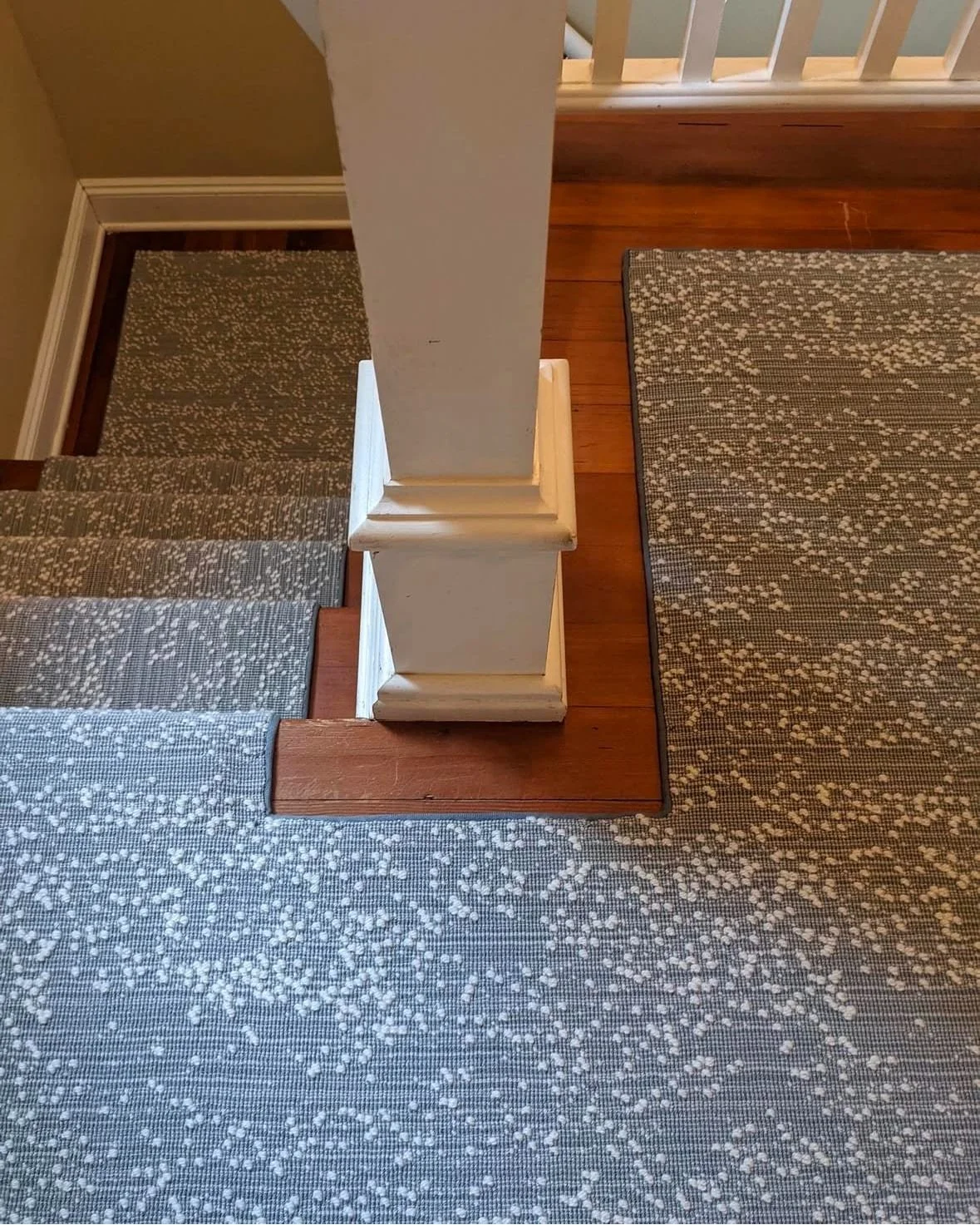 Top-down view of a staircase with a white painted wooden post, wooden steps, and inlaid carpets on the stairs and floor.