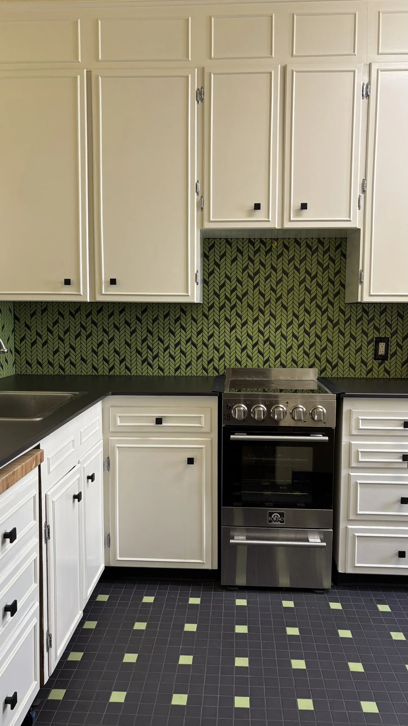 Kitchen with white cabinets, black countertop, and a patterned green backsplash. Contains a gas stove and black square drawer and cabinet knobs.
