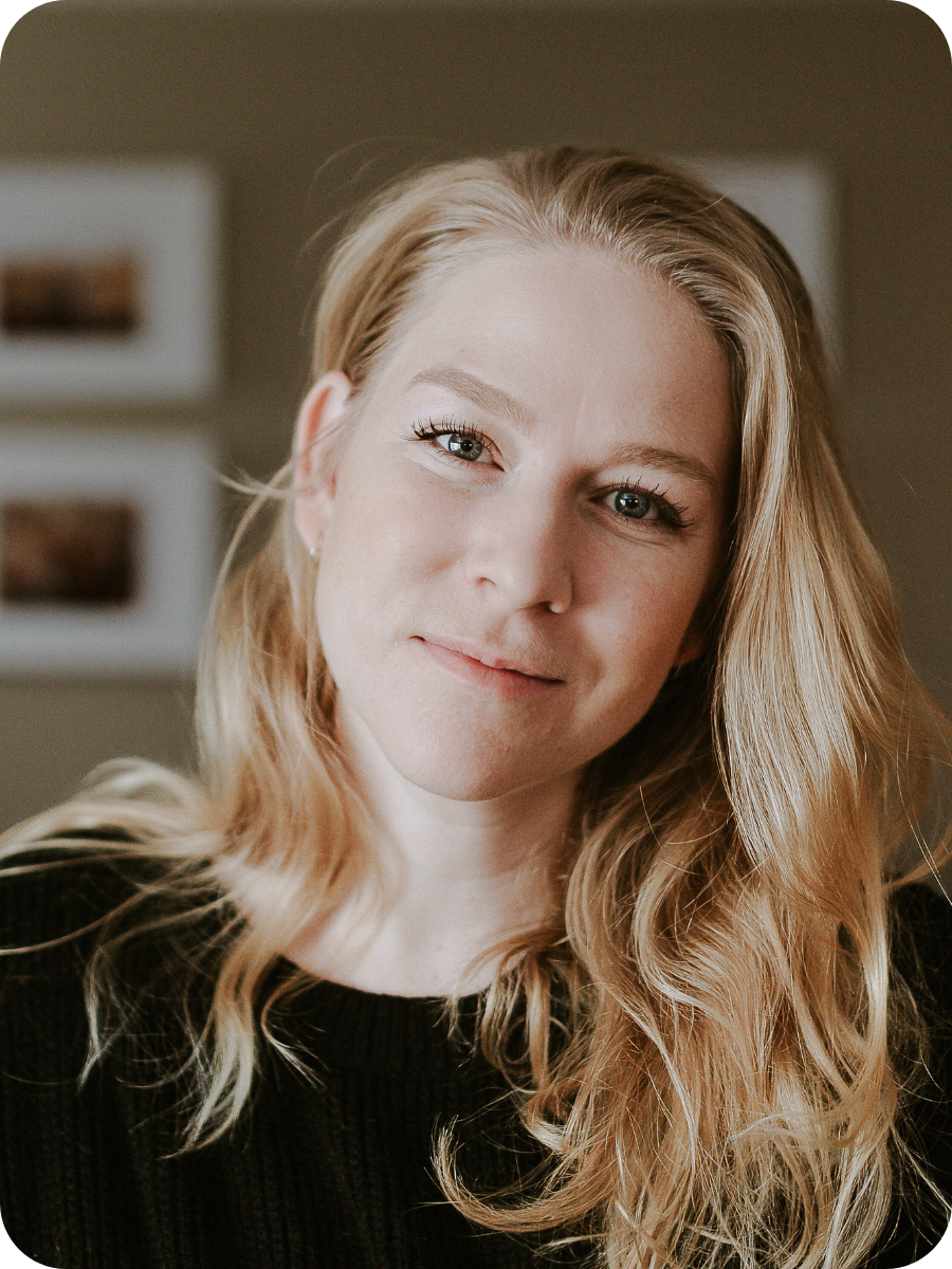 Close-up portrait of a young woman with blonde hair, blue eyes, and a slight smile, wearing a black top, indoors with a blurred background.