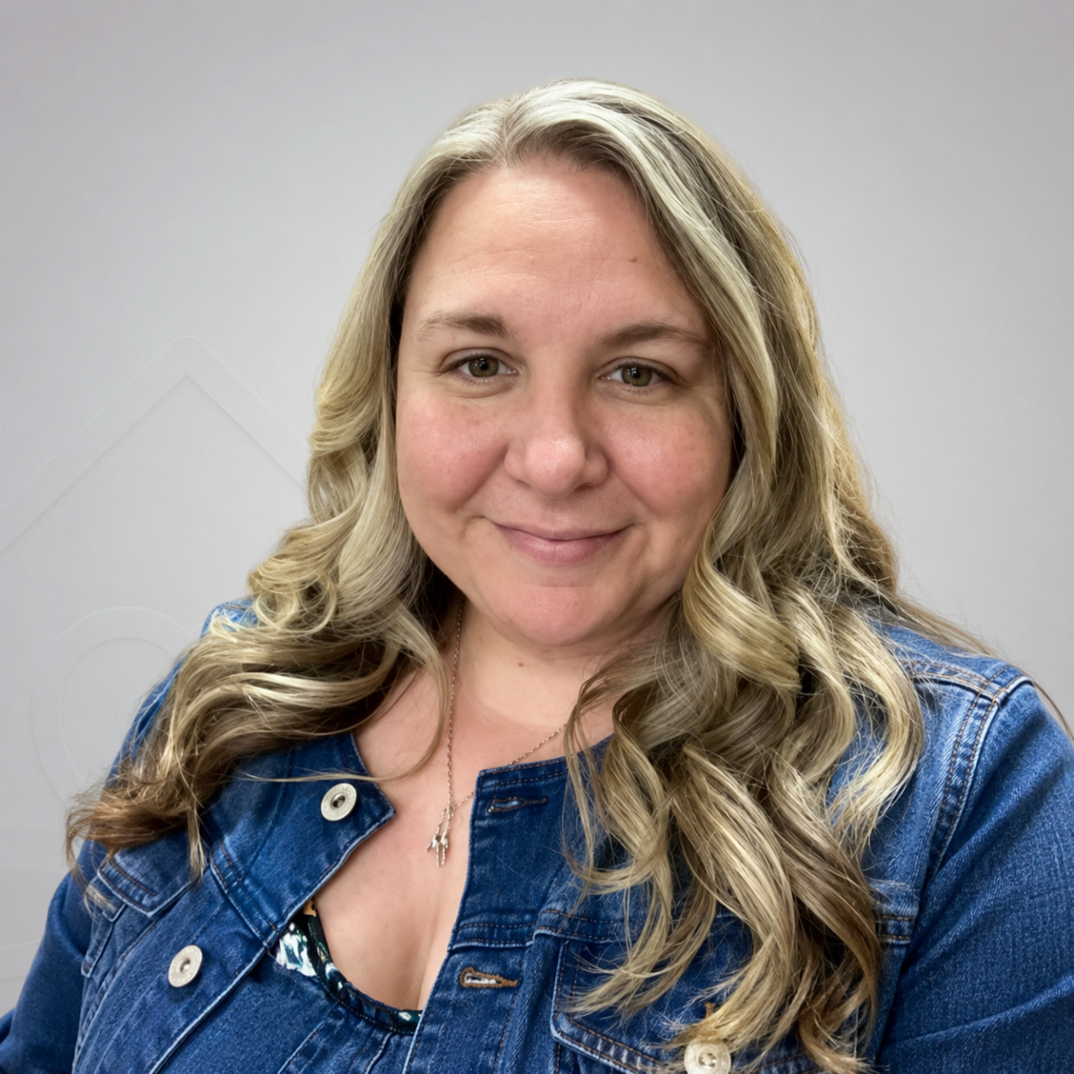 A woman with long, blonde, wavy hair, wearing a denim jacket and a necklace, smiling at the camera against a black background.