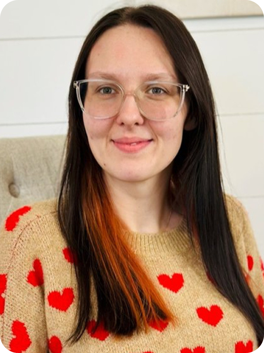 A woman with long dark hair and glasses smiling, wearing a beige sweater with red hearts, sitting in a light-colored chair indoors.