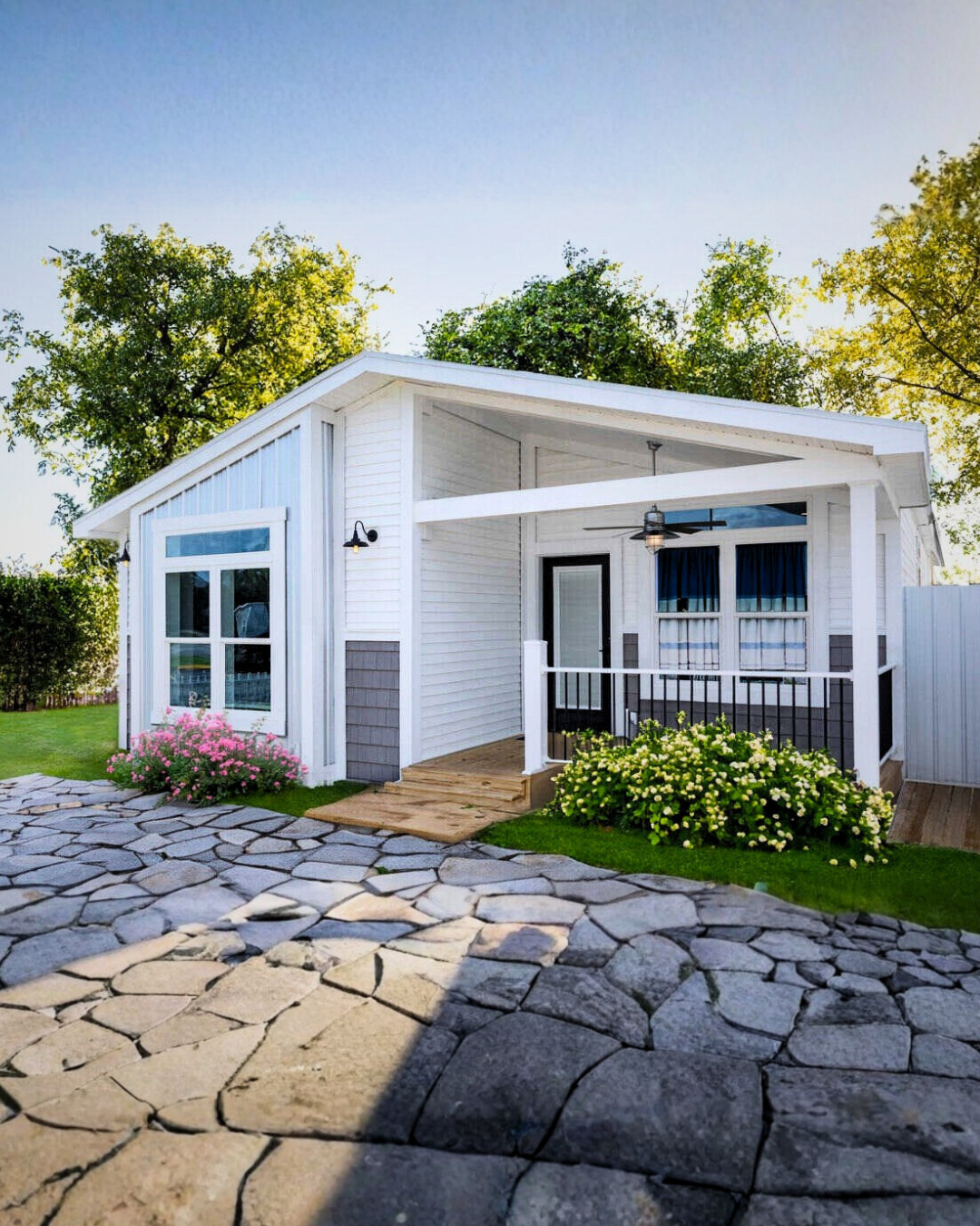 A modern white house with a small porch, black railings, and a glass door, surrounded by green grass and bushes with pink and white flowers, under a clear blue sky.