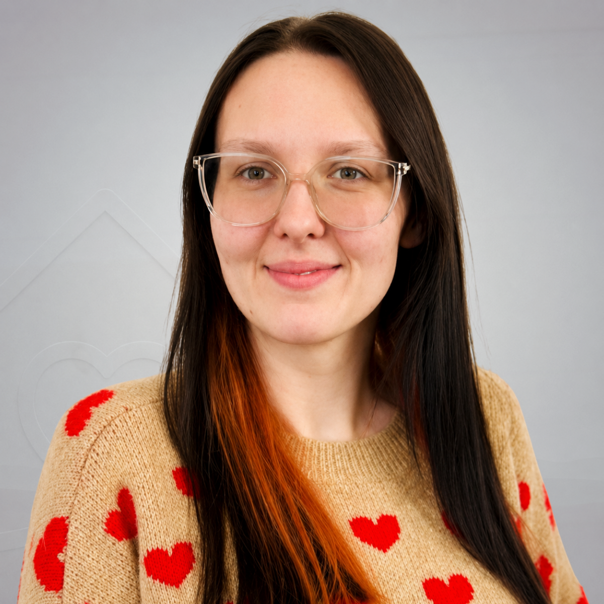 A woman with long dark hair and glasses smiling, wearing a beige sweater with red hearts, sitting in a light-colored chair indoors.