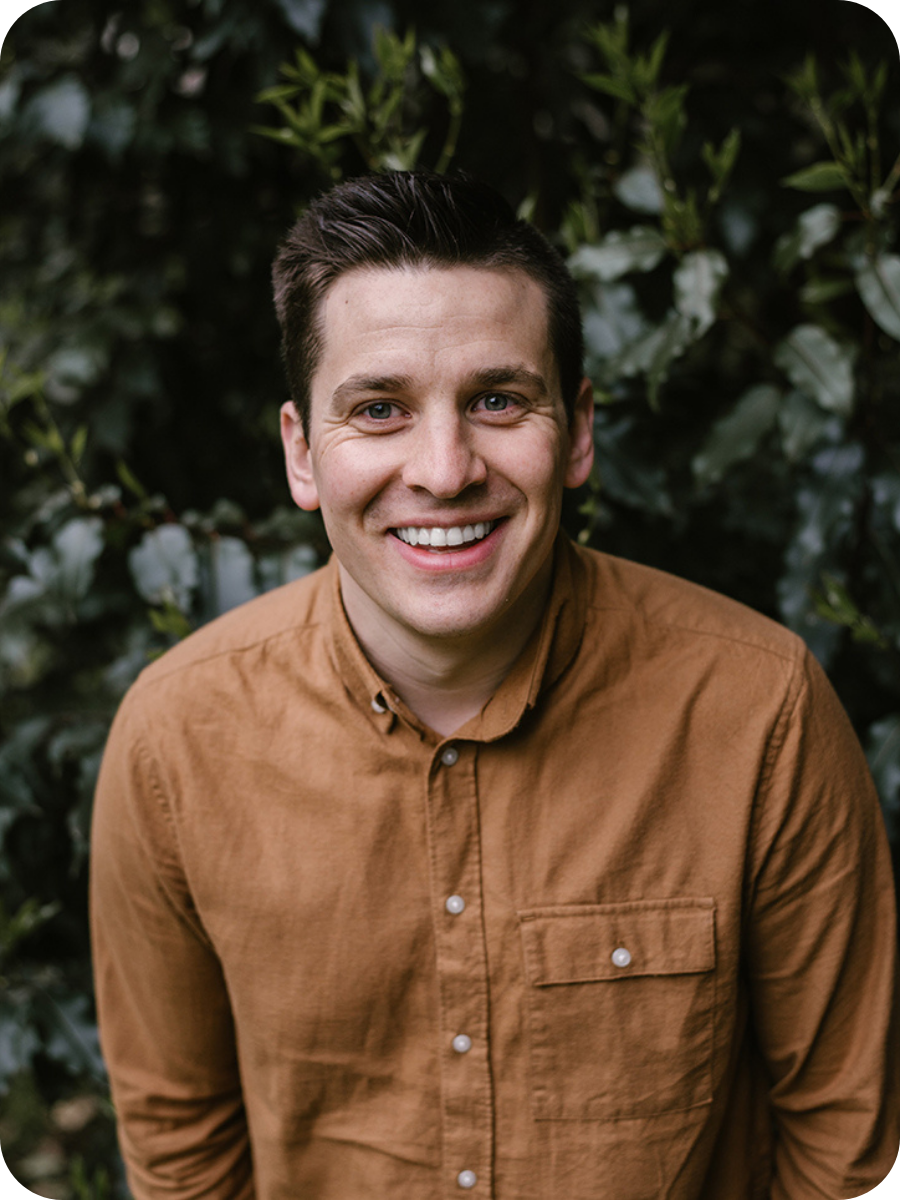 A man with dark hair and a beard, wearing an orange button up, standing in front of green trees