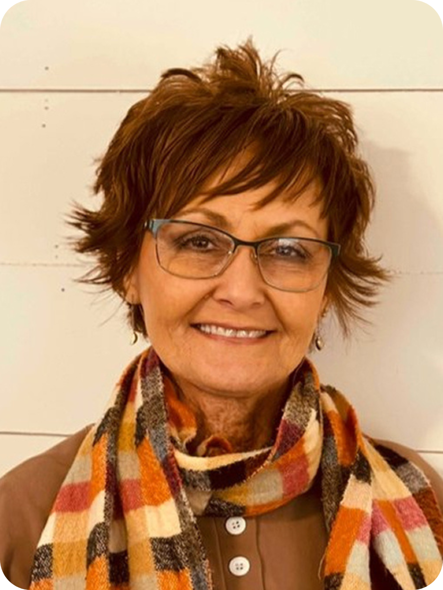 A smiling woman with short, layered brown hair and glasses, wearing a multicolored plaid scarf and a brown top, standing against a white wall.