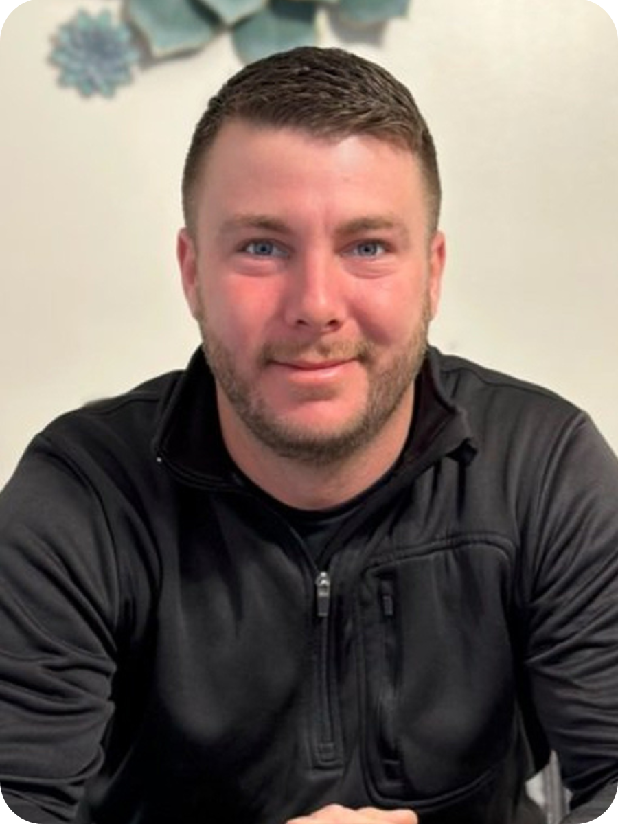 A man with short brown hair and a beard, wearing a black athletic jacket, sitting indoors with a light-colored wall and some decorative flower or snowflake cutouts in the background.