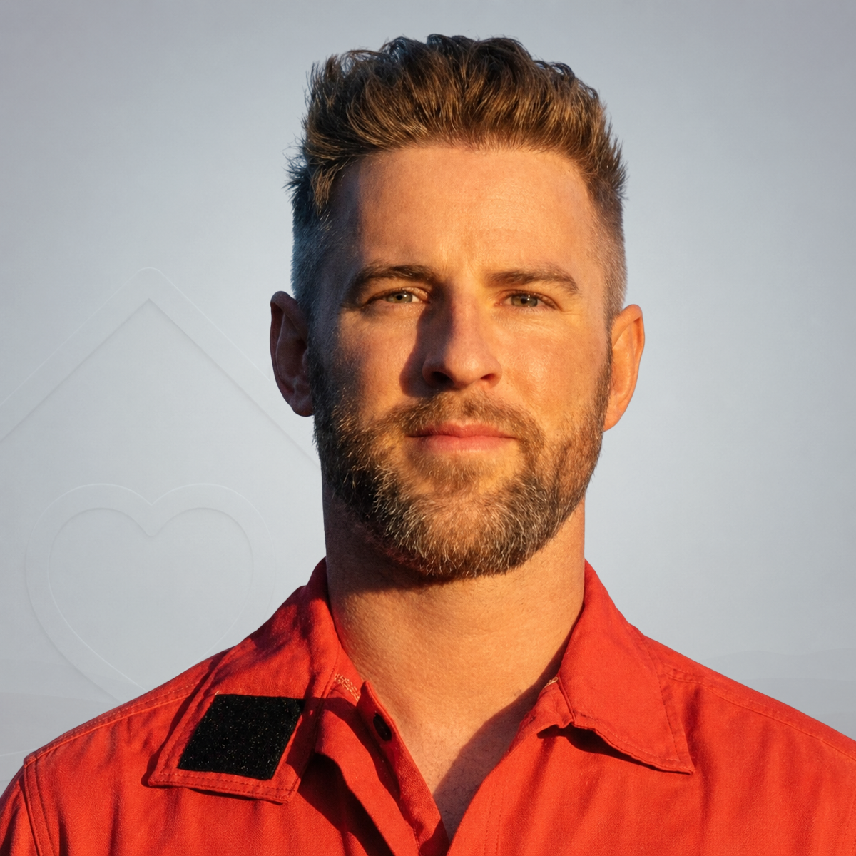 A man with short, styled hair and a beard wearing an orange or red shirt against a blue sky background.