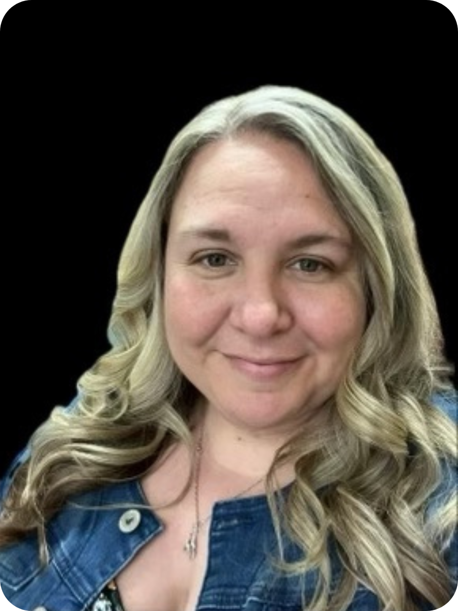 A woman with long, blonde, wavy hair, wearing a denim jacket and a necklace, smiling at the camera against a black background.