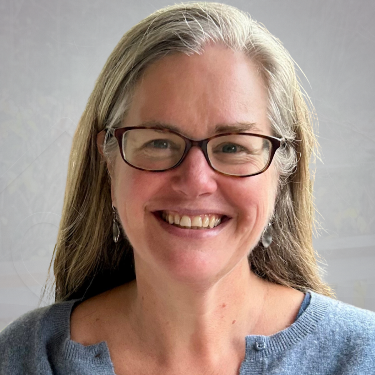 A woman with gray hair, glasses, and earrings smiling outdoors in front of trees and a wooden fence.