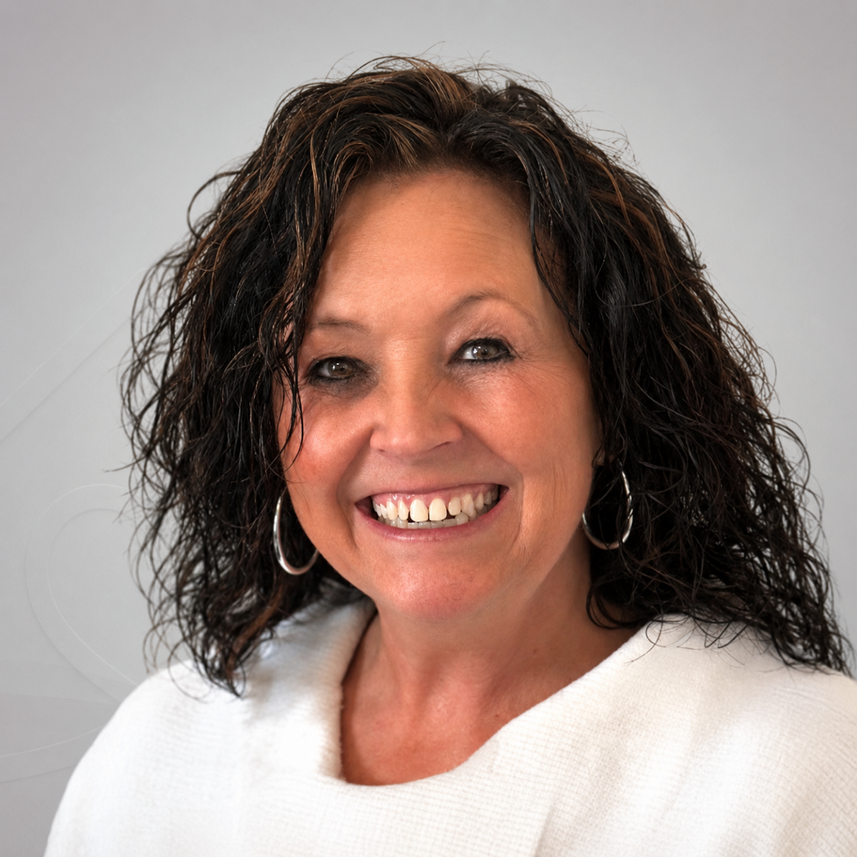 Smiling woman with curly dark hair, hoop earrings, and a light-colored top against a plain background.