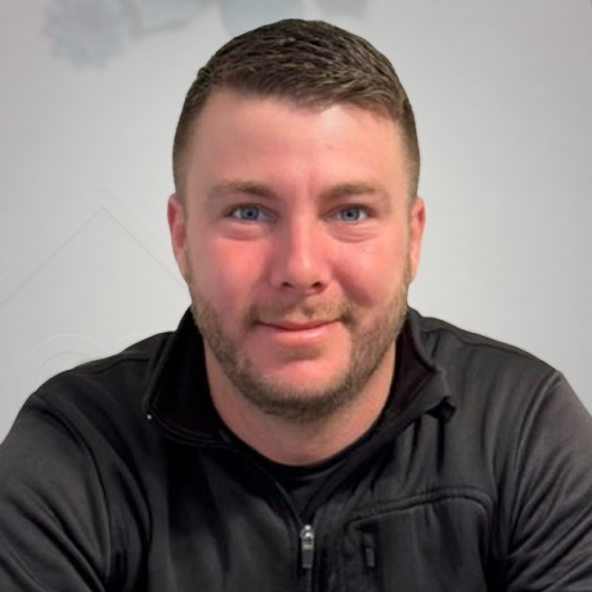A man with short brown hair and a beard, wearing a black athletic jacket, sitting indoors with a light-colored wall and some decorative flower or snowflake cutouts in the background.