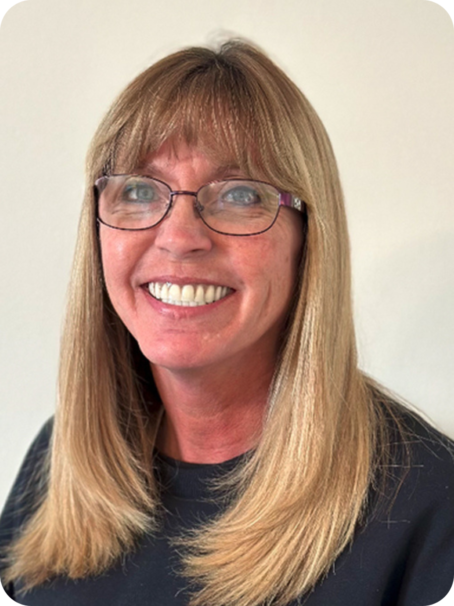 Smiling woman with long blonde hair, glasses, and a black top against a plain light-colored background.