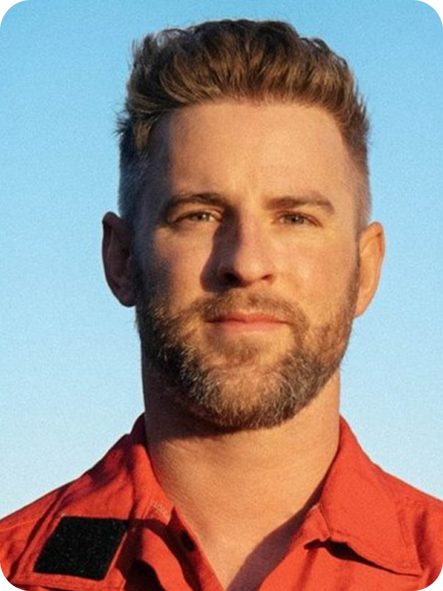 A man with short, styled hair and a beard wearing an orange or red shirt against a blue sky background.