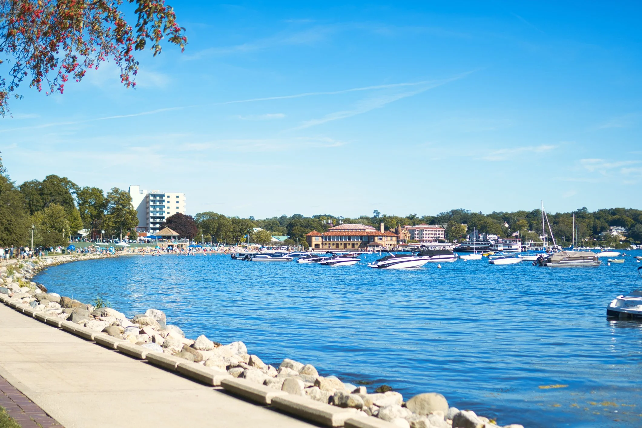 A lakeside scene with a river or lake, lined with rocks and a walking path. Several boats are docked on the water, and there are trees and buildings in the background under a clear blue sky.