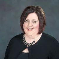 Portrait of a woman with dark bob hairstyle, wearing a black blazer and a beaded necklace, smiling against a gray background.